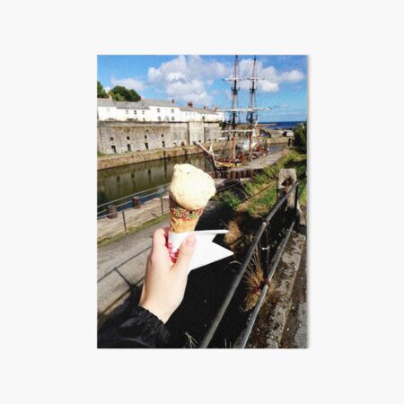 Person holding an ice cream cone near a sailboat and an old building, sunny day with clouds in the sky Art Board Print