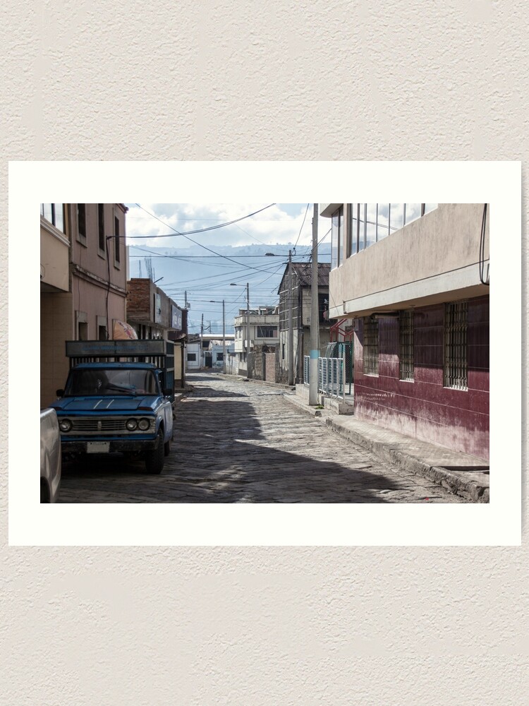 Alternate view of Empty quiet winding cobblestone street in Guamote, Ecuador with blue truck, red tile building, power lines and street lamps Art Print
