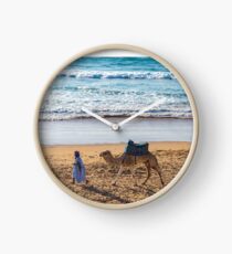 Reloj Hombre caminando un camello mientras otro reza en Taghazout, Marruecos.