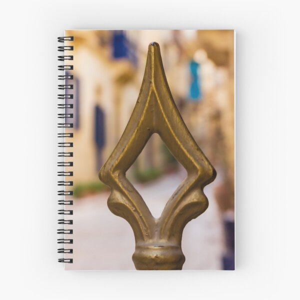 Artikelvorschau, Nahaufnahme einer Messing-Zierspitze an einem verzierten Zaun mit sanftem Straßenbokeh und warmen mediterranen Farbtönen entworfen und verkauft von Nmallia.