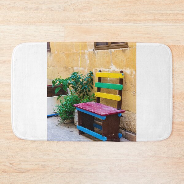 Item preview, Colorful Wooden Bench Against Textured Yellow Wall in Sunny Courtyard With Green Plants designed and sold by Nmallia.