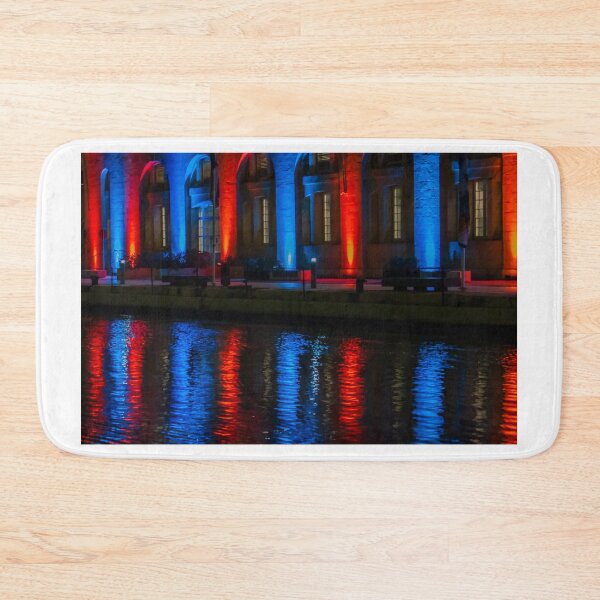 Item preview, Nighttime Waterfront Building With Red And Blue Illuminated Arches And Reflections On Water designed and sold by Nmallia.