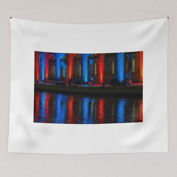 Item preview, Nighttime Waterfront Building With Red And Blue Illuminated Arches And Reflections On Water designed and sold by Nmallia.