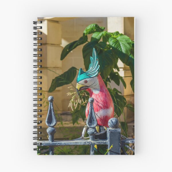 Item preview, Colorful Painted Bird Statue Perched on a Weathered Metal Fence Amid Lush Greenery designed and sold by Nmallia.