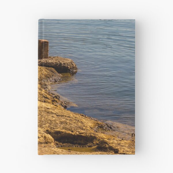 Item preview, Rugged Coastal Rocks By Calm Blue Water With Weathered Stone Pier And Small Post designed and sold by Nmallia.