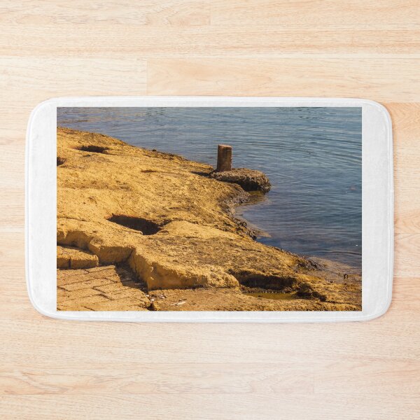 Item preview, Rugged Coastal Rocks By Calm Blue Water With Weathered Stone Pier And Small Post designed and sold by Nmallia.